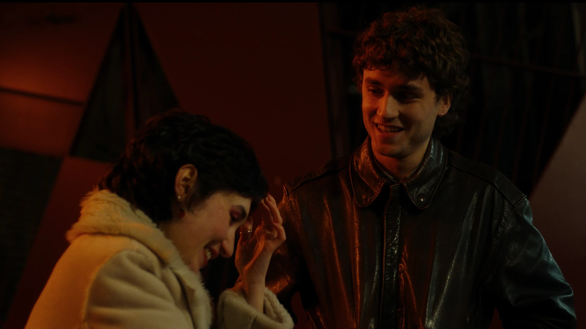 Still from Cool About It: A young woman and young man laugh outside a bar with red lights overhead.
