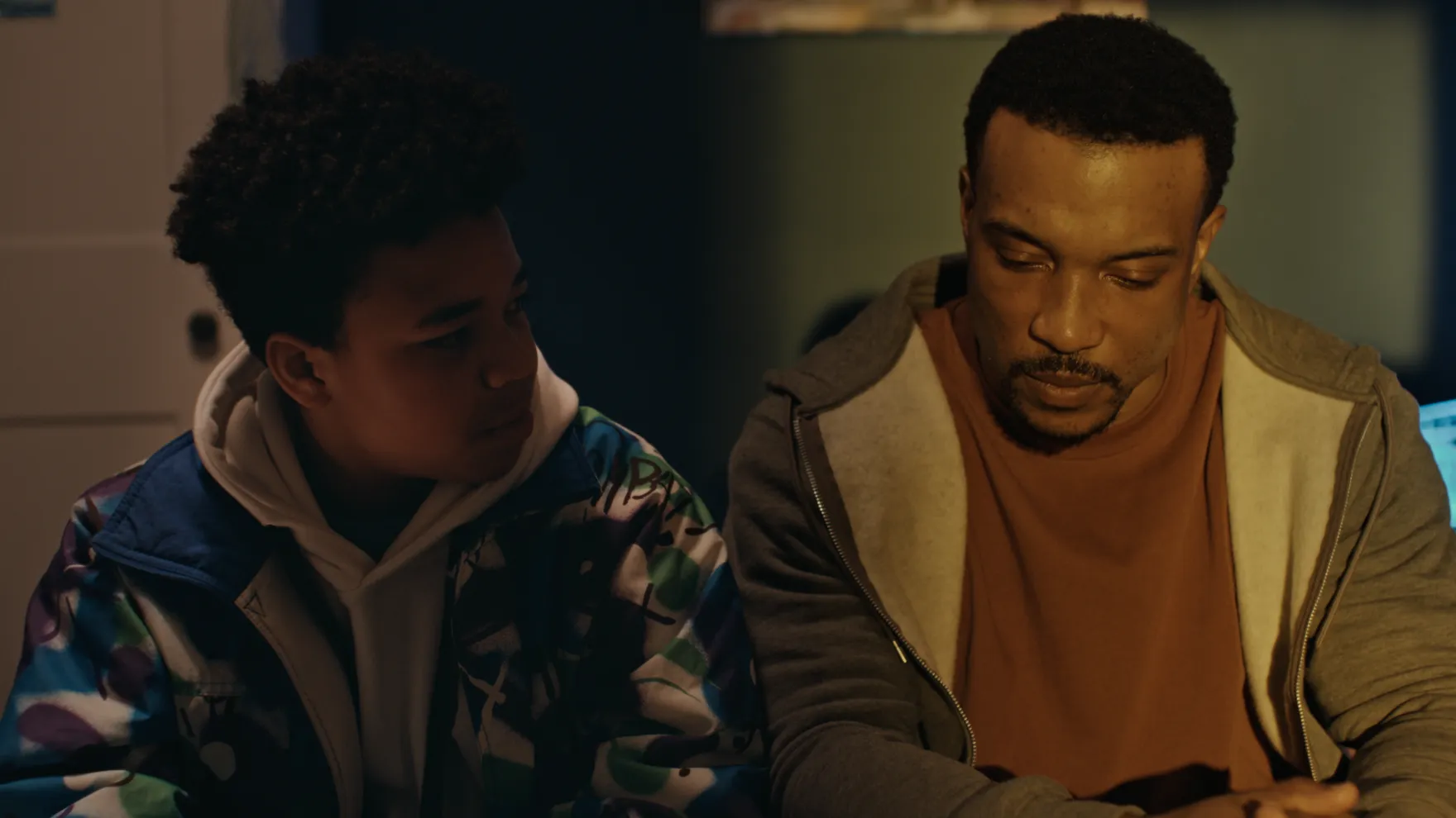 A boy in a colourful patterned jacket sits beside a man in a hoodie and t-shirt, who looks down with a serious expression in a dimly lit room.