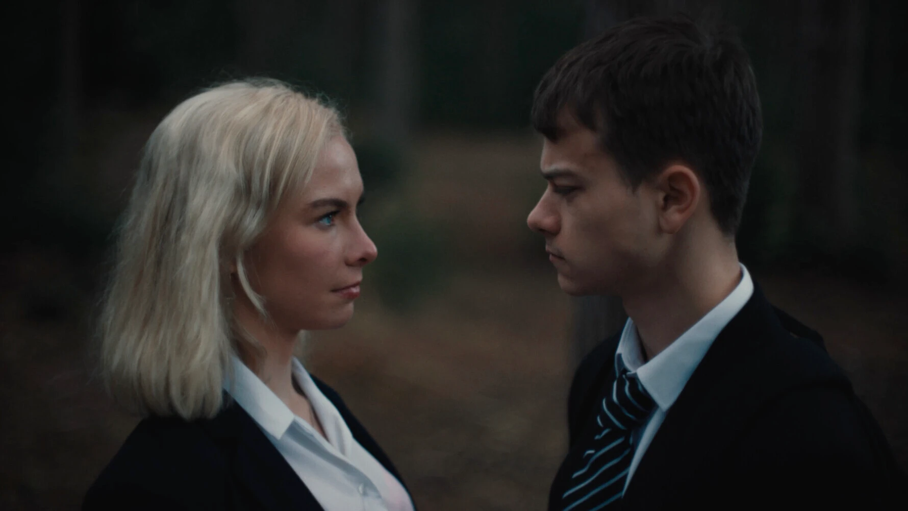 Two students in school uniforms facing each other outdoors, blonde girl and dark-haired boy having conversation