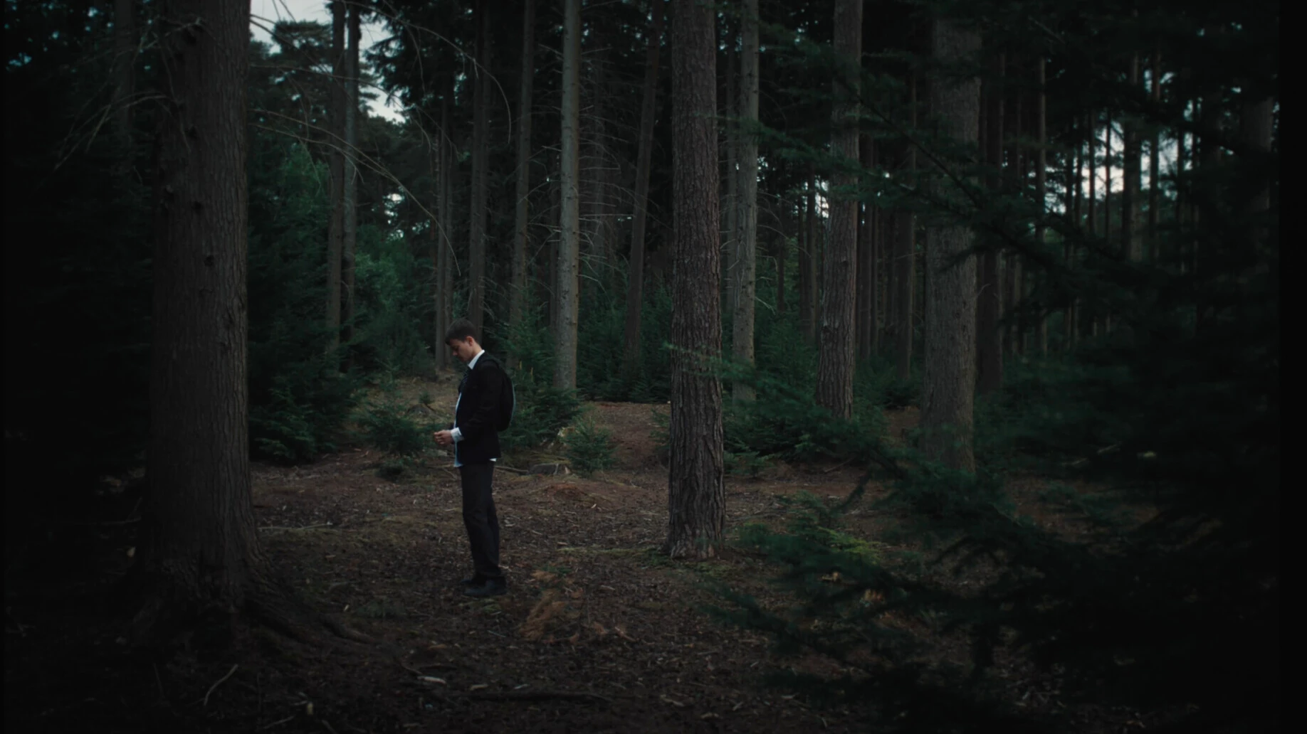 Man in black school uniform standing alone in dense forest with tall trees at dawn
