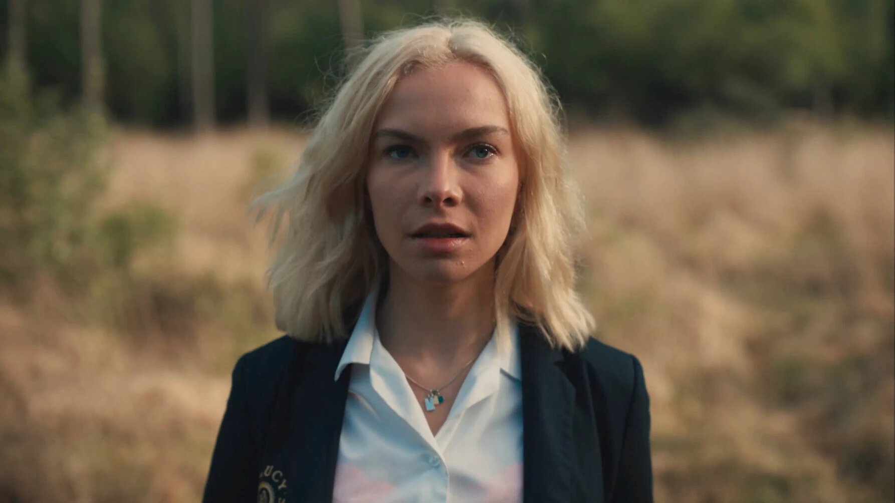 Young woman with blonde hair wearing school uniform with blazer and white shirt standing outdoors in natural setting