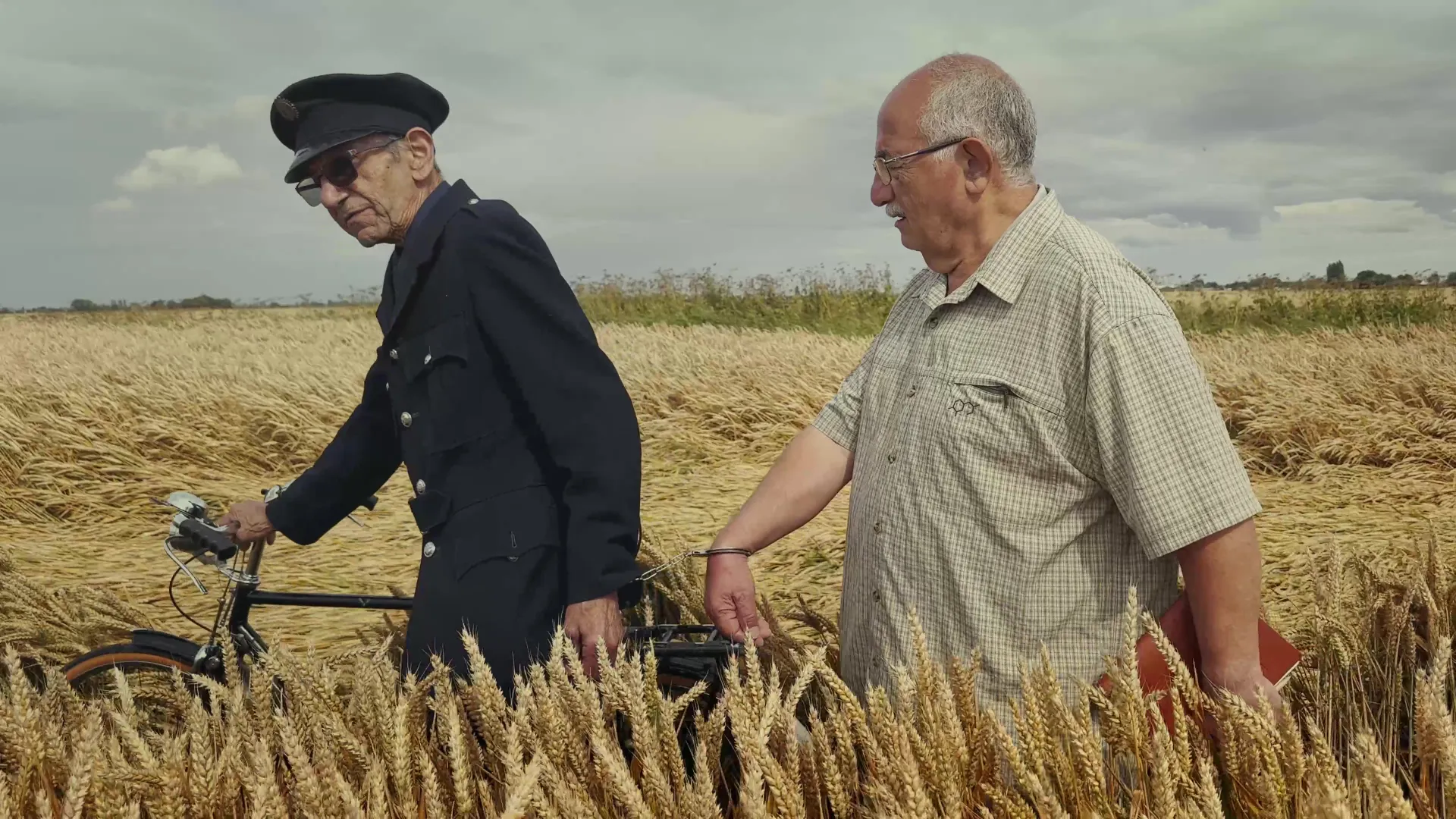 An elderly policeman and an old man, chained together, walk through a wheat field as the policeman carries his vintage bicycle.