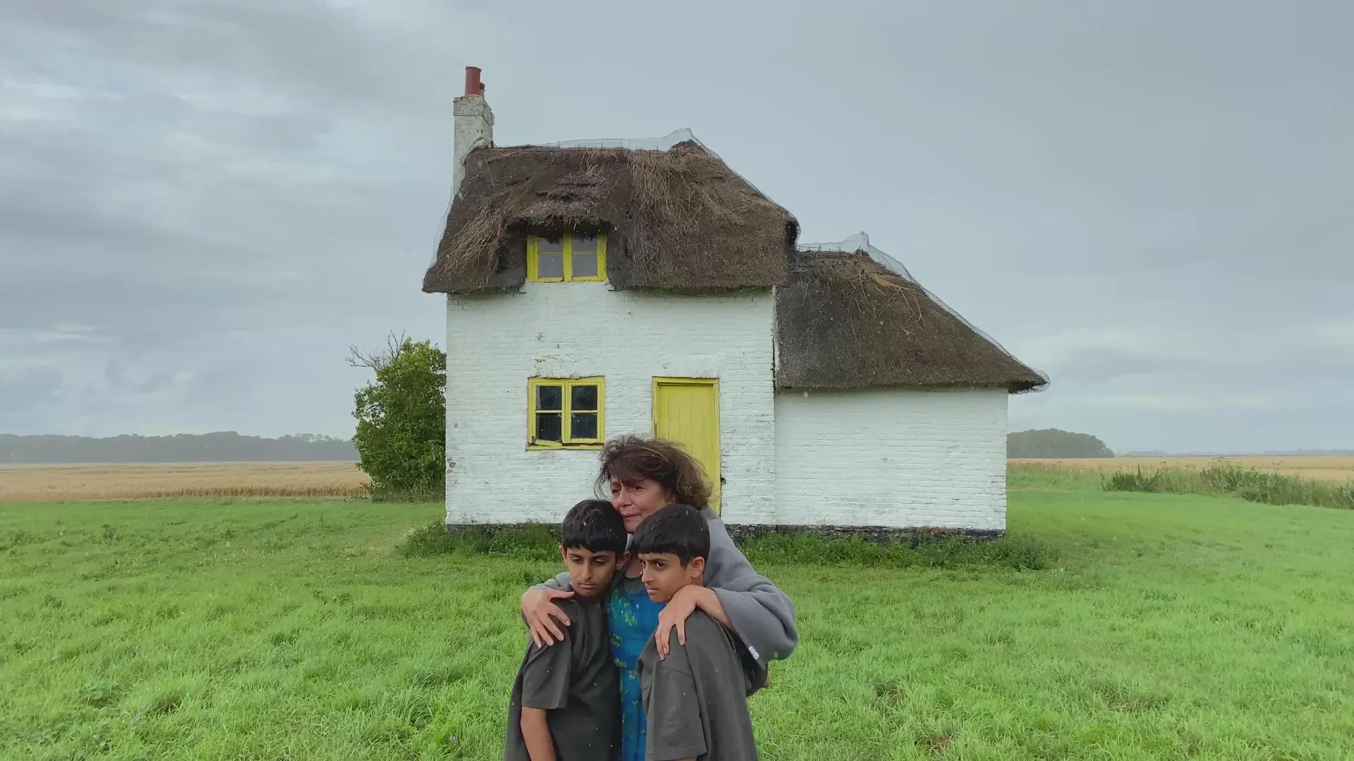 A woman stands in front of a small white cottage with a thatched roof and yellow windows, gently embracing two boys on a green field under a cloudy sky.