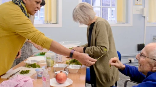 A lady passes a herb to an elderly man, while an elderly woman prepares food behind.