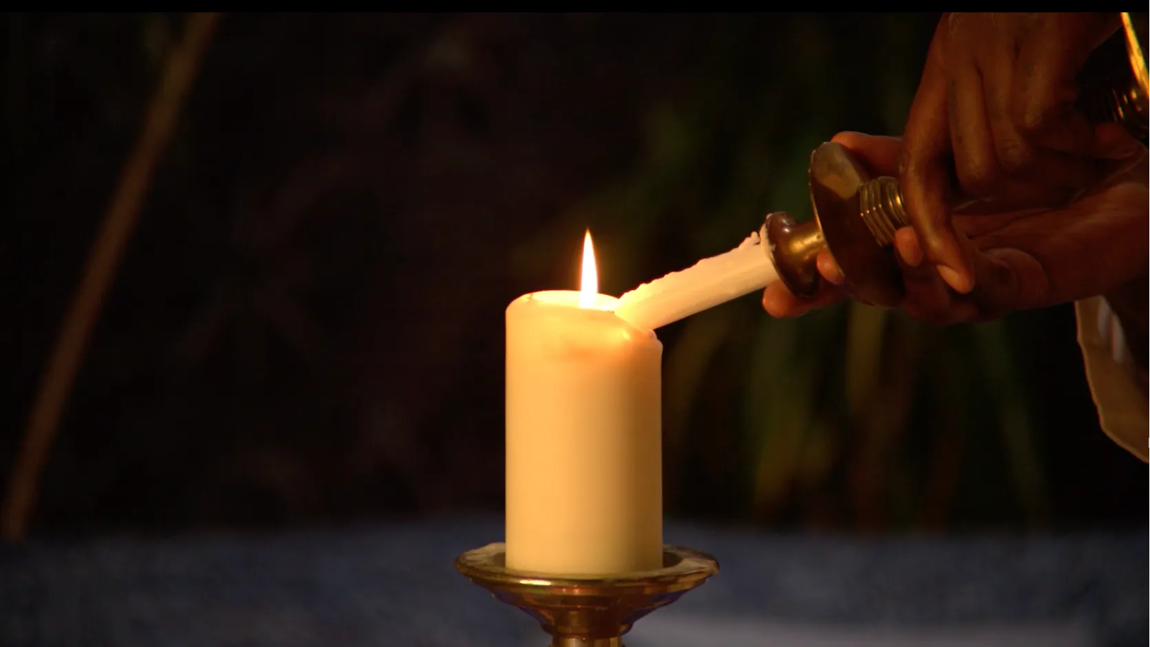 A hand holding a lit taper candle lights a larger pillar candle placed on a metal candle holder, with a dark blurred background.