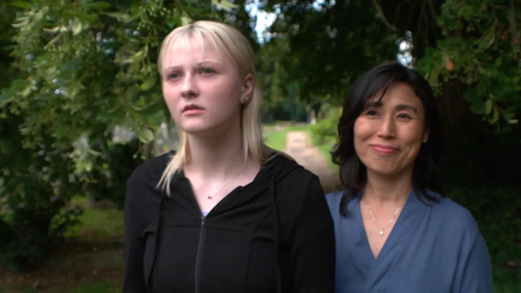Two women, one younger and one older, standing in a cemetery, looking beyond the camera