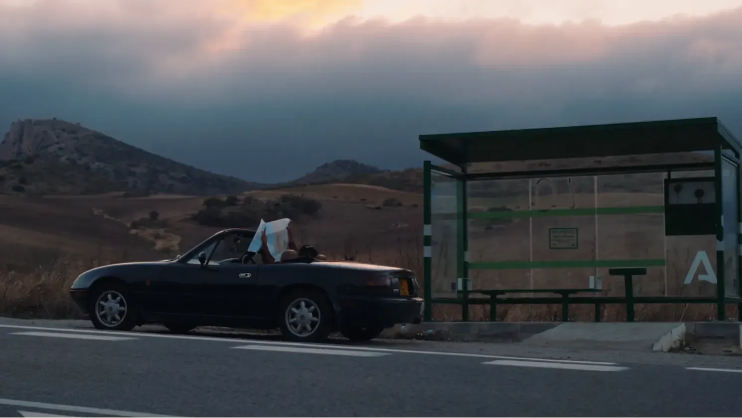 Hills and a moody sky are the backdrop to someone studying a map in an open-topped car, parked by a bus shelter.