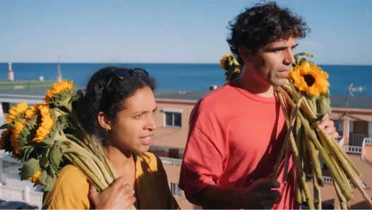 A woman and a man in the sunshine by the sea, both clutching several sunflowers