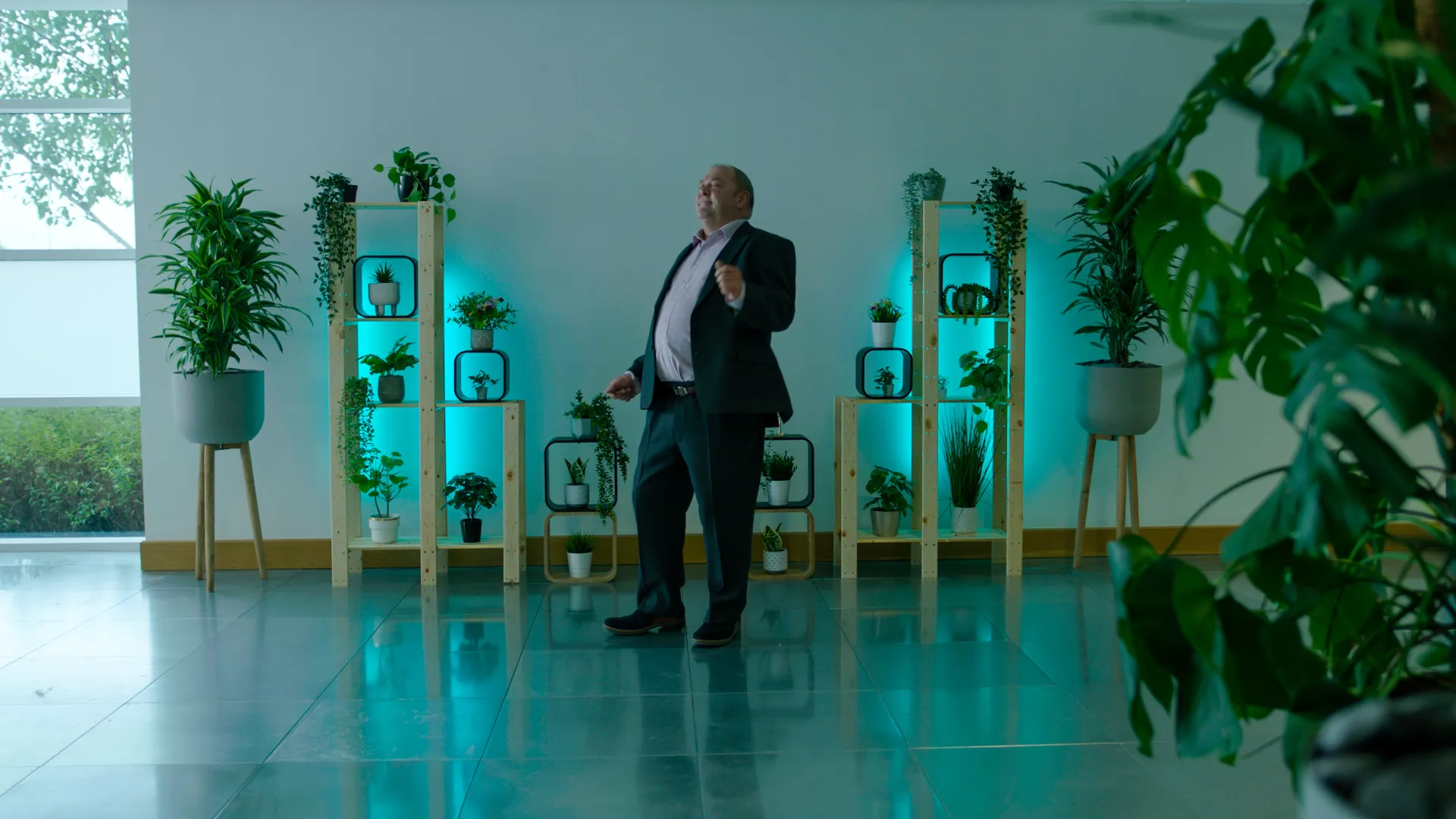 A middle-aged man in a grey suit dances awkwardly before flowers, green light reflecting off his blue suede shoes in a cold, clinical room.