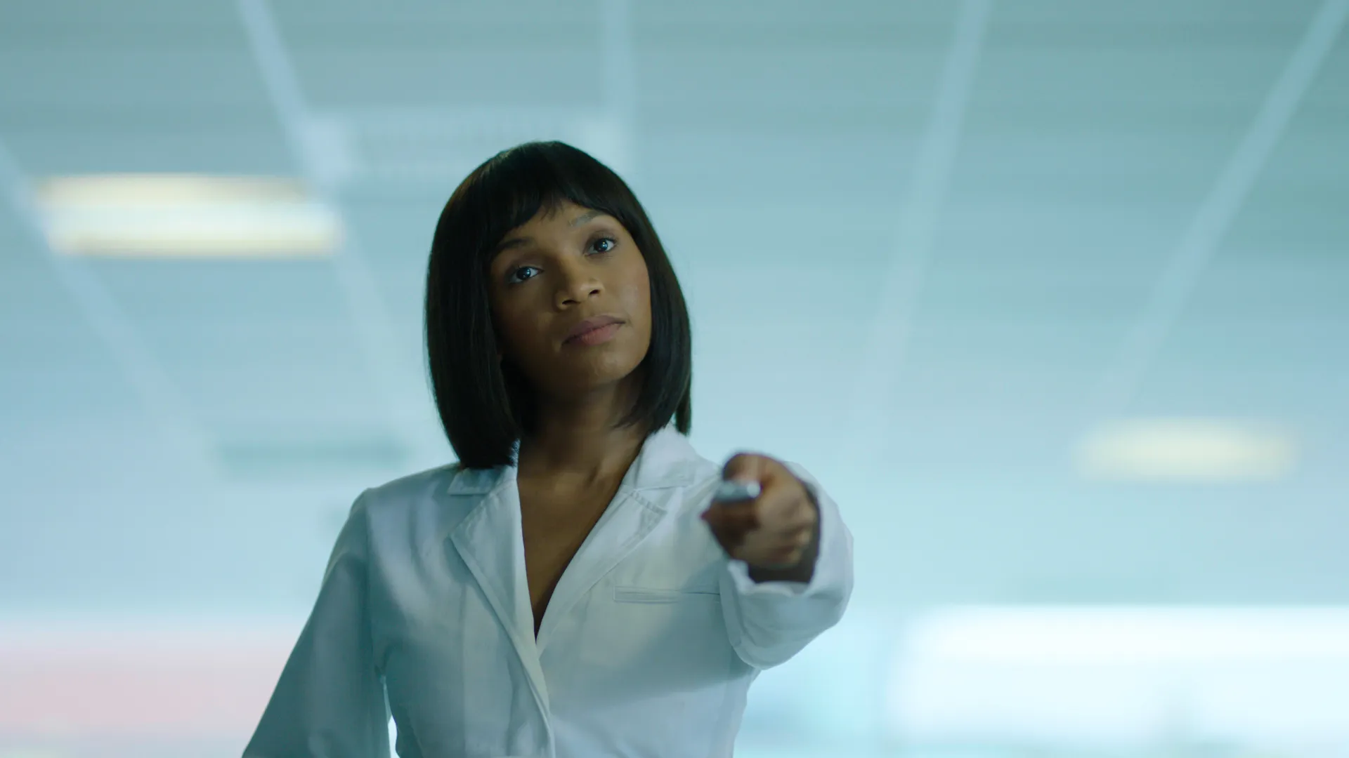 A young Black woman in a white doctor’s coat extends a remote in a sterile, white-lit room glowing with cold, dystopian light.