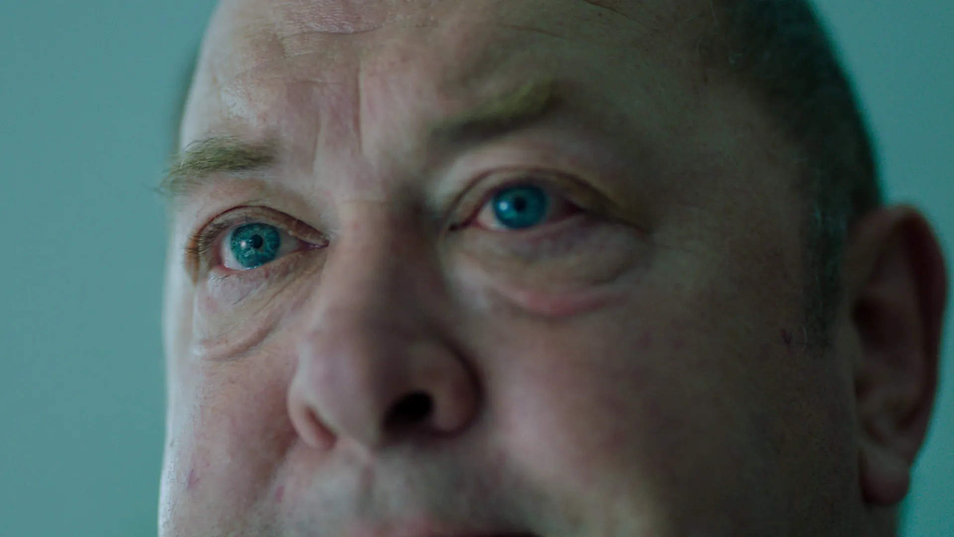 A weary white man in his sixties stares ahead, blue eyes rimmed red, as mint-green light casts a cold, dystopian glow around him.