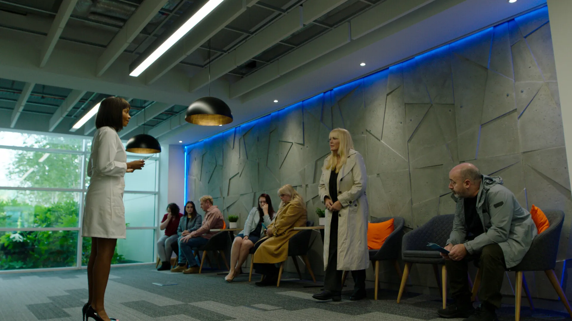 In a grey, sterile waiting room, eight weary people sit silently as a Black woman in a white coat stands before them amid a cold, dystopian glow.