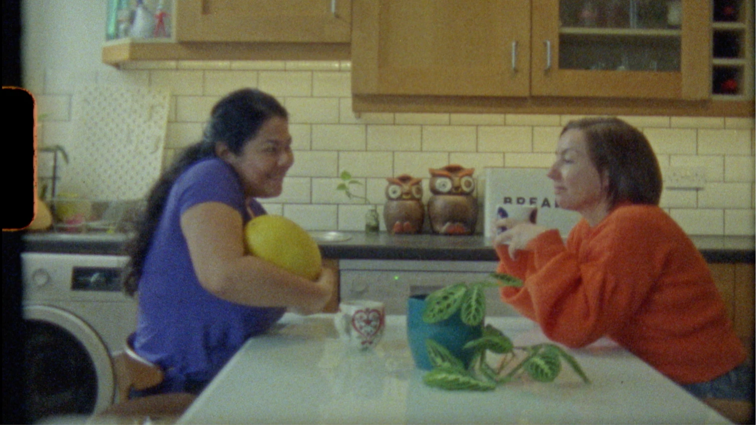 A woman in a purple top holding a melon and a woman in an orange top holding a coffee mug sit at a kitchen table