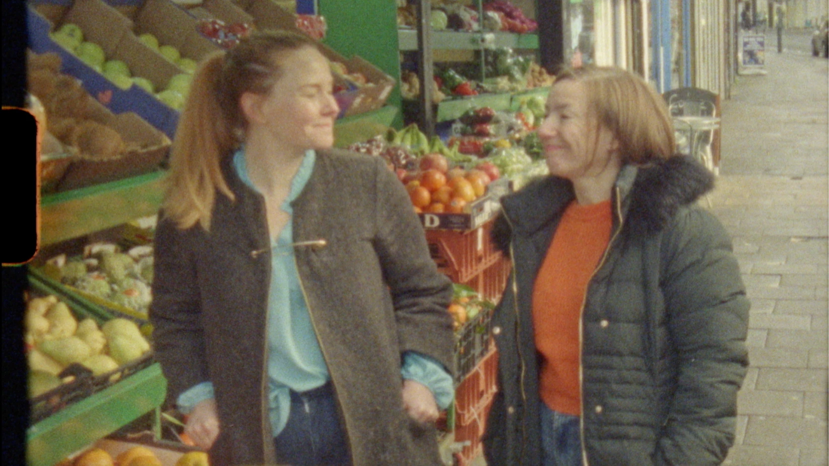 A woman in a grey coat and a woman in a black coat smile at each other on the pavement outside a fruit stand
