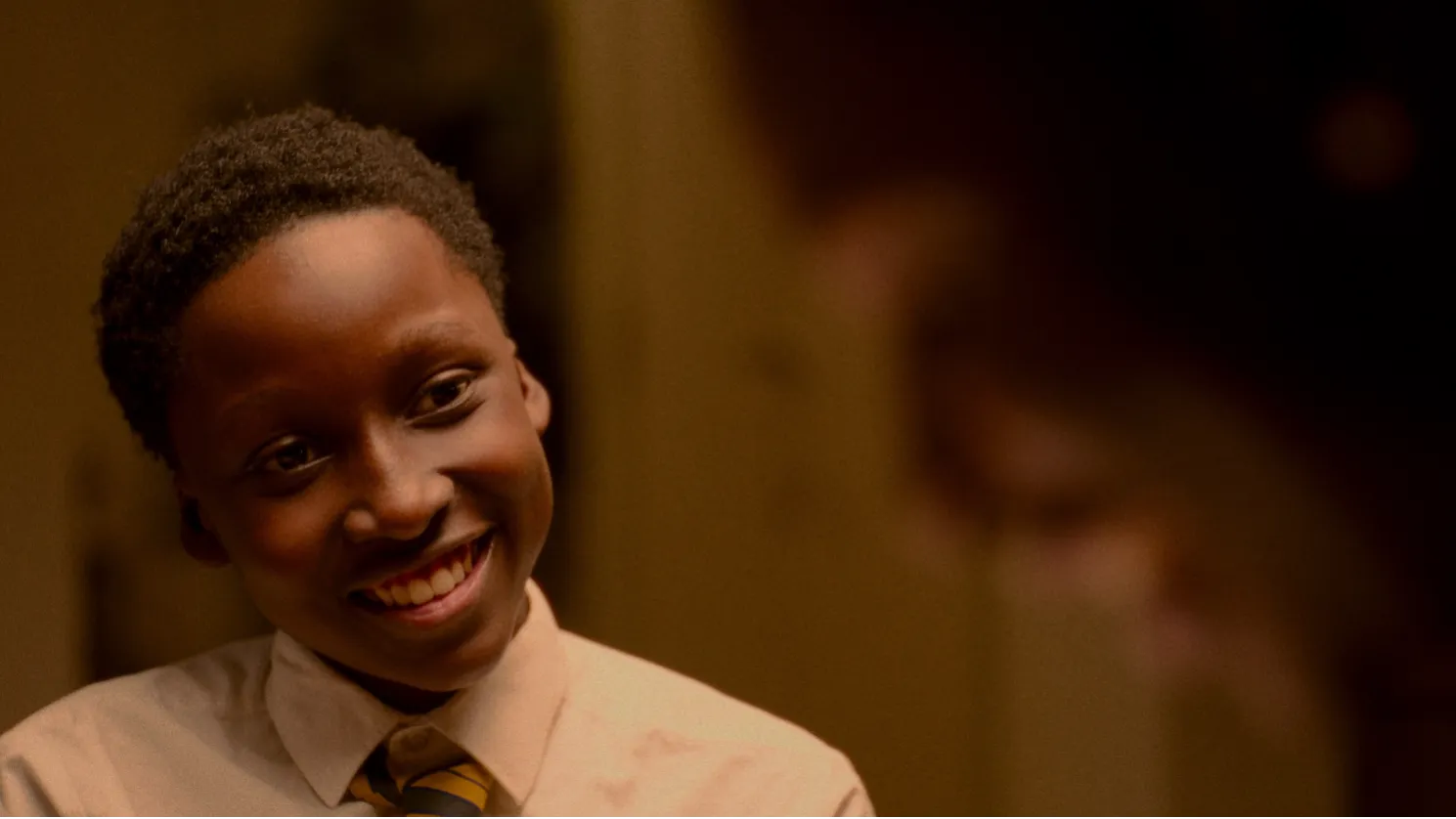 A young Black boy wearing school uniform grins at someone, who is seen out of focus at the edge of the image frame.