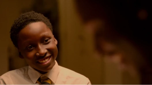 A young Black boy wearing school uniform grins at someone, who is seen out of focus at the edge of the image frame.