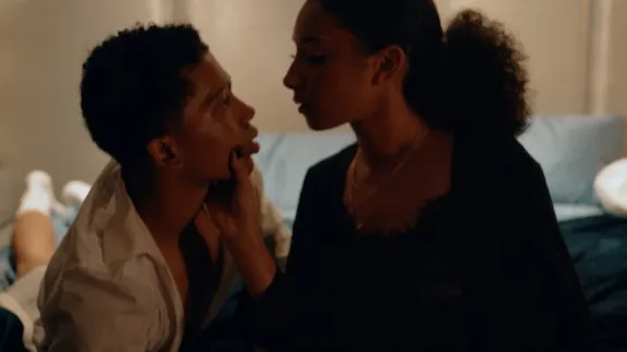 A woman and a young boy are sitting on a bed. The woman holds the boy's face in a way that shows control while he stares at her as if under a spell.