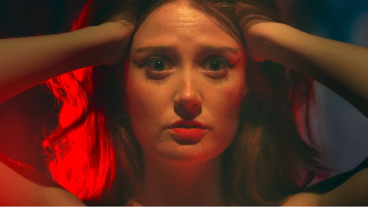Red-haired performer clutching her head in dramatic lighting, illuminated by intense red and blue hues, staring wide-eyed in a tense cinematic close-up.