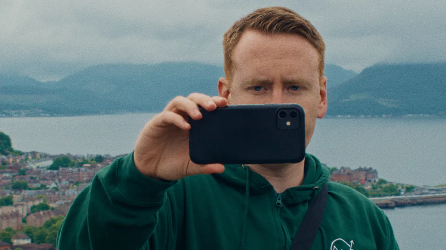 Outdoors. A man holds a mobile phone in front of him and records using the phone's camera.