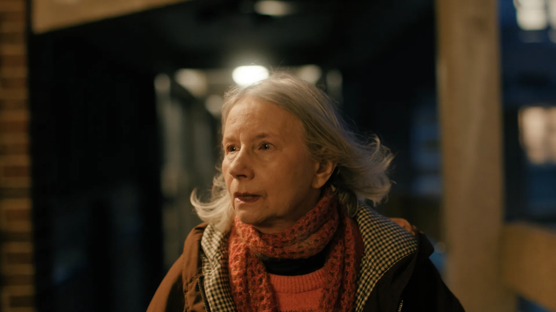 A medium close up of an older woman facing camera walking in an outdoor corridor at night