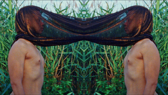 A man faces a mirror image of himself - the two men are joined by a brown sheet material