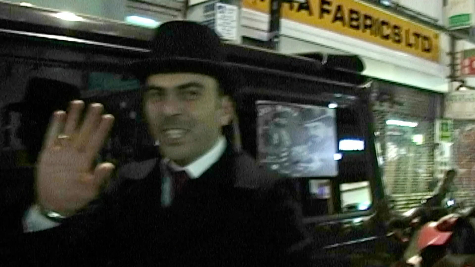 A man in formal dark clothing and a hat standing beside a black vehicle at night, raising his hand toward the camera on a busy city street lined with lit shop signs.