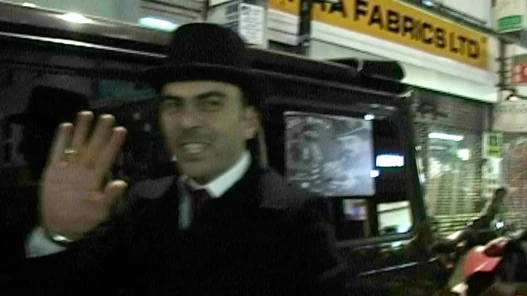 A man in formal dark clothing and a hat standing beside a black vehicle at night, raising his hand toward the camera on a busy city street lined with lit shop signs.