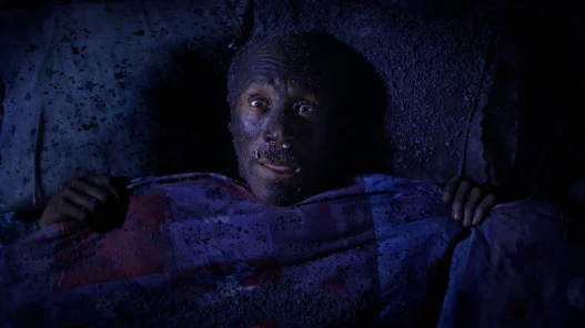 Danny John-Jules as Deen Peels in Sound Asleep, lies in a burned out bed, eyes wide, facing camera.