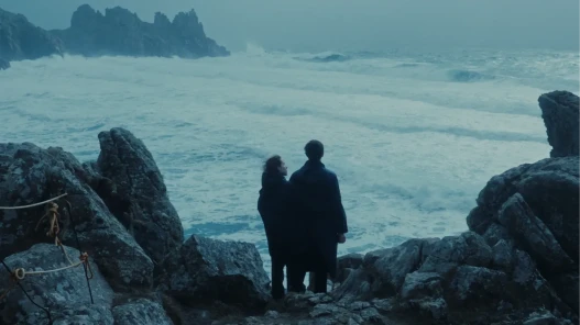 A woman and a man stand on a stormy cliff edge looking out to sea.