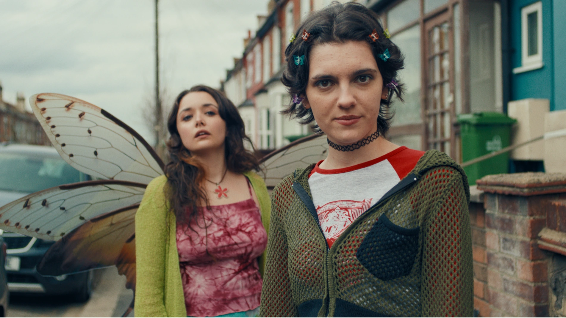 Stood on a street are two sisters, one is looking into the camera and the other is looking away and has giant moth wings. They are dressed in 2000s fashion.