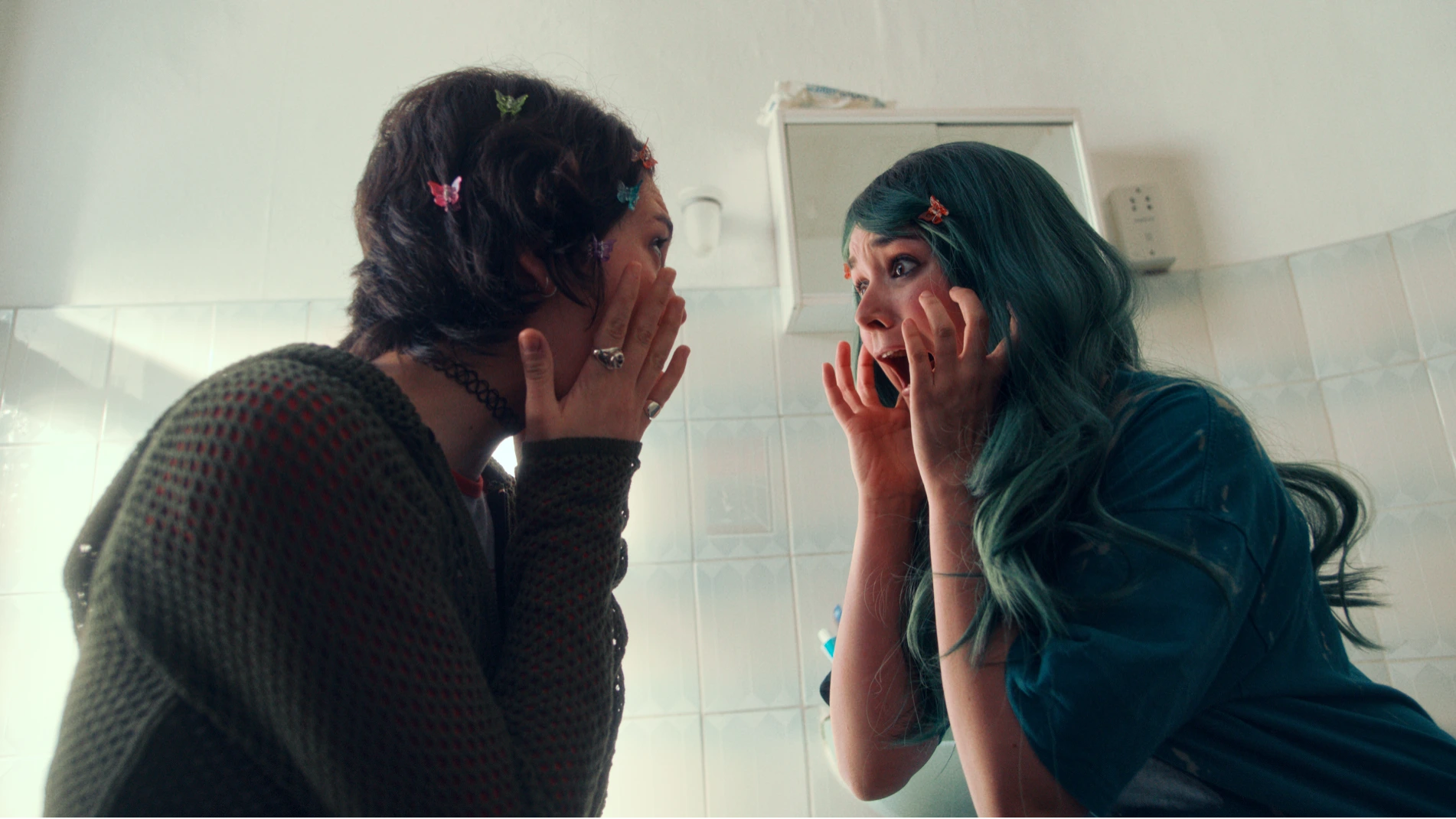 Two sisters are in a bathroom and looking at each other and screaming with their hands on their faces. One of the sisters has blue hair.