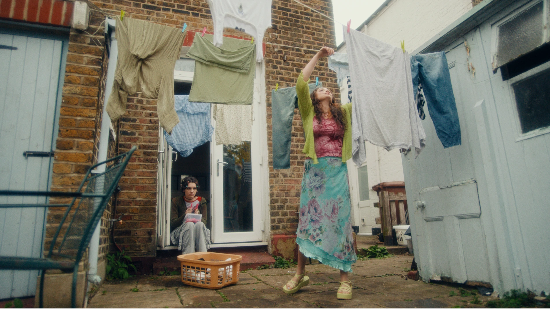 Two sisters are in their backyard. One if hanging up washing and the other is looking bored sat on the step.