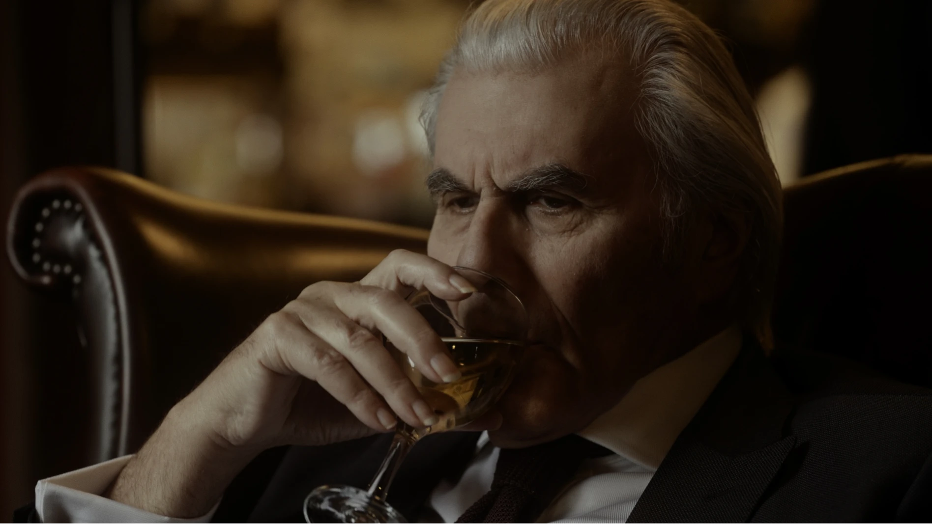 A smartly dressed man sat in an armchair drinking a glass of white wine