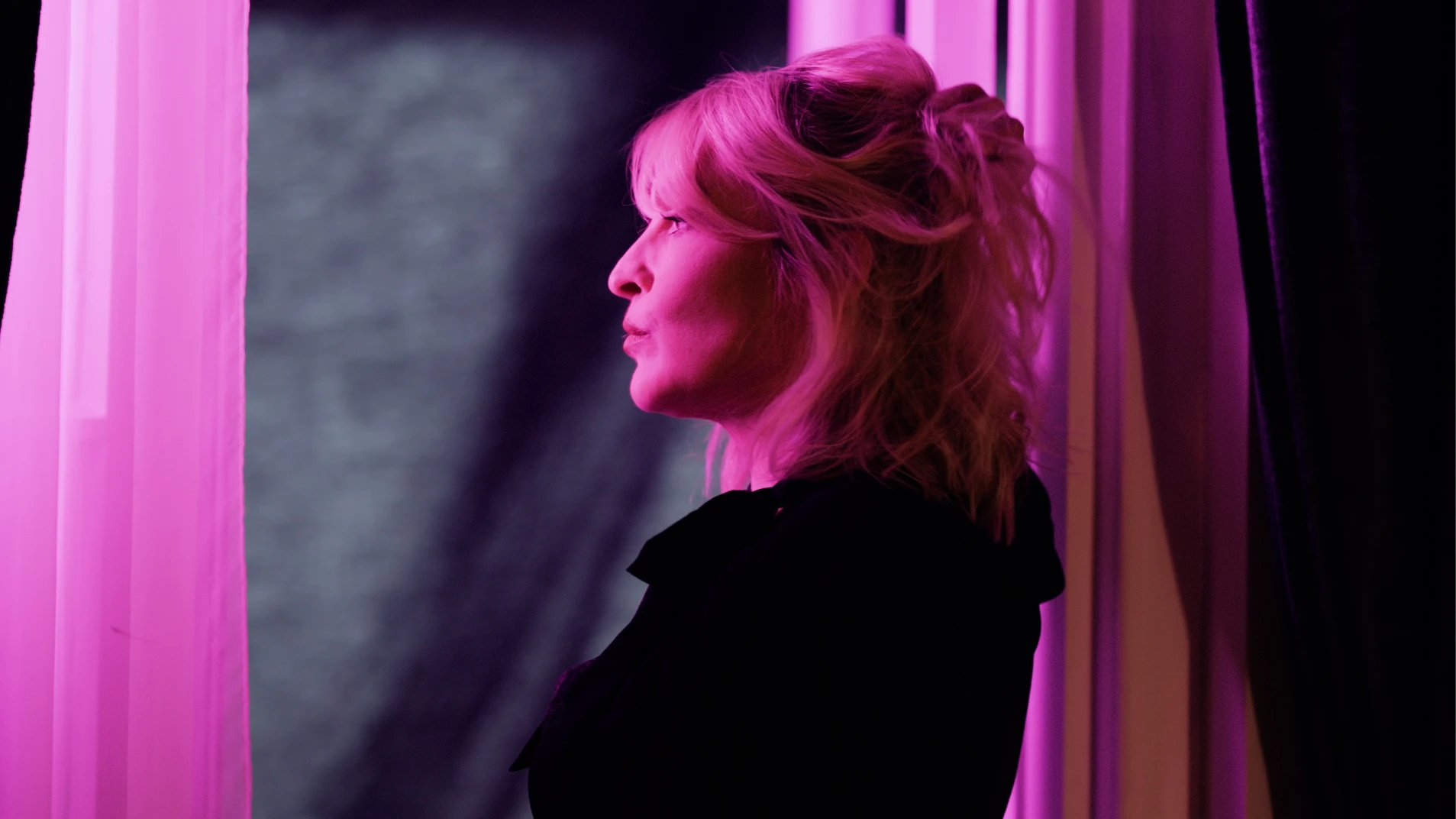 A woman staring out the window at night bathed in neon pink light