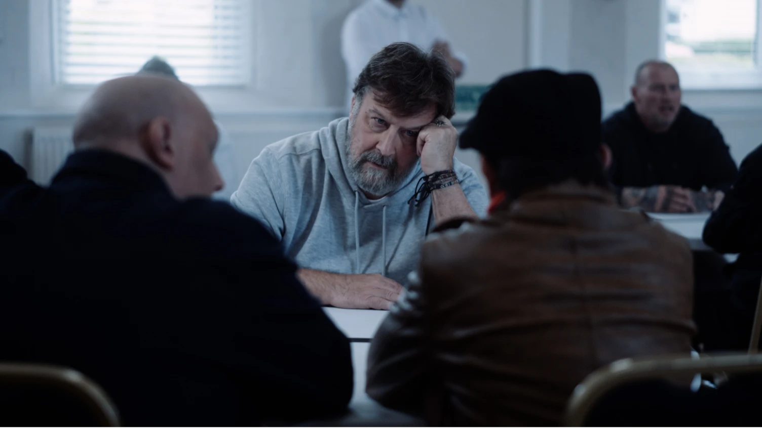 Three men sat around the table in a prison visiting room with other prisoners and guards visible in the background