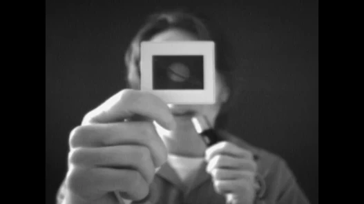 Black and white close up of woman holding a film slide of planet Saturn in front of face while holding microphone to mouth