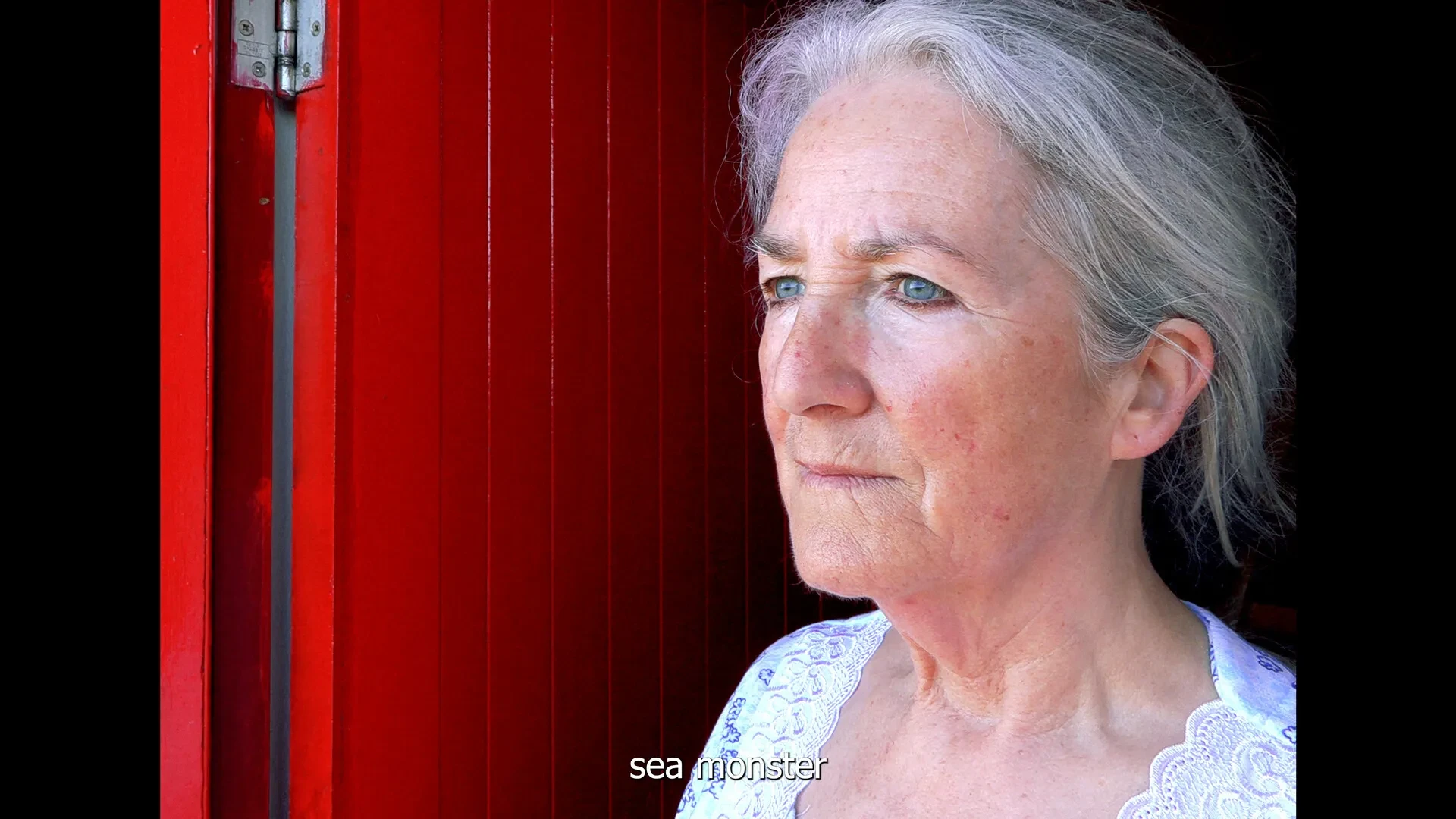 A woman with grey hair and blue eyes looking out from a red door