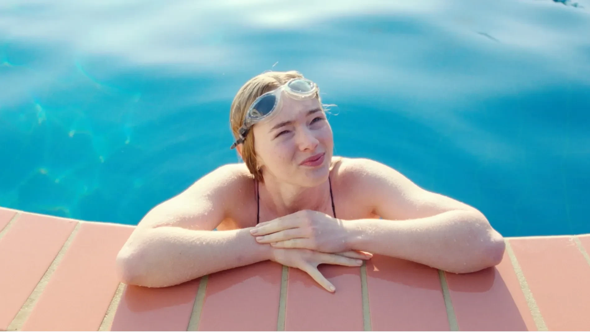 A young woman leans on the side of the pool smiling.