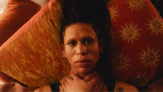 A mixed race woman looks directly into the camera with her hands around her own neck, lying against orange velvet pillows.