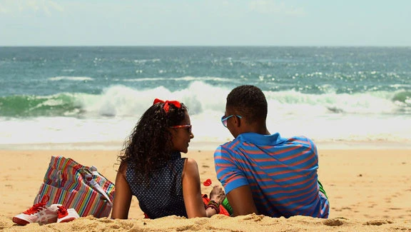 Young couple sit closely together on a sandy beach, the girl holding a heart-shaped lollipop, framed as a cinematic still with vibrant colour.