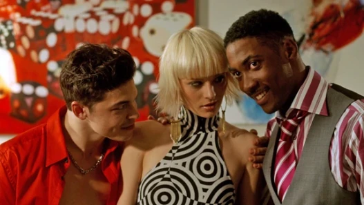 Two young men and a young woman stand indoors, the woman centred, dressed in high-end fashion, posing with composed confidence, with a casino-style dice board in the background