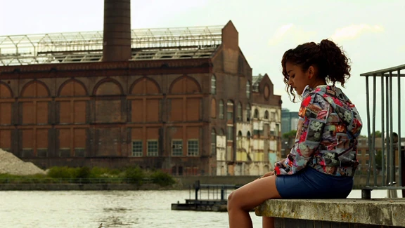 Teenage girl in a hooded jacket sits alone on a London river barge, with the Lots Road Power Station in the background.
