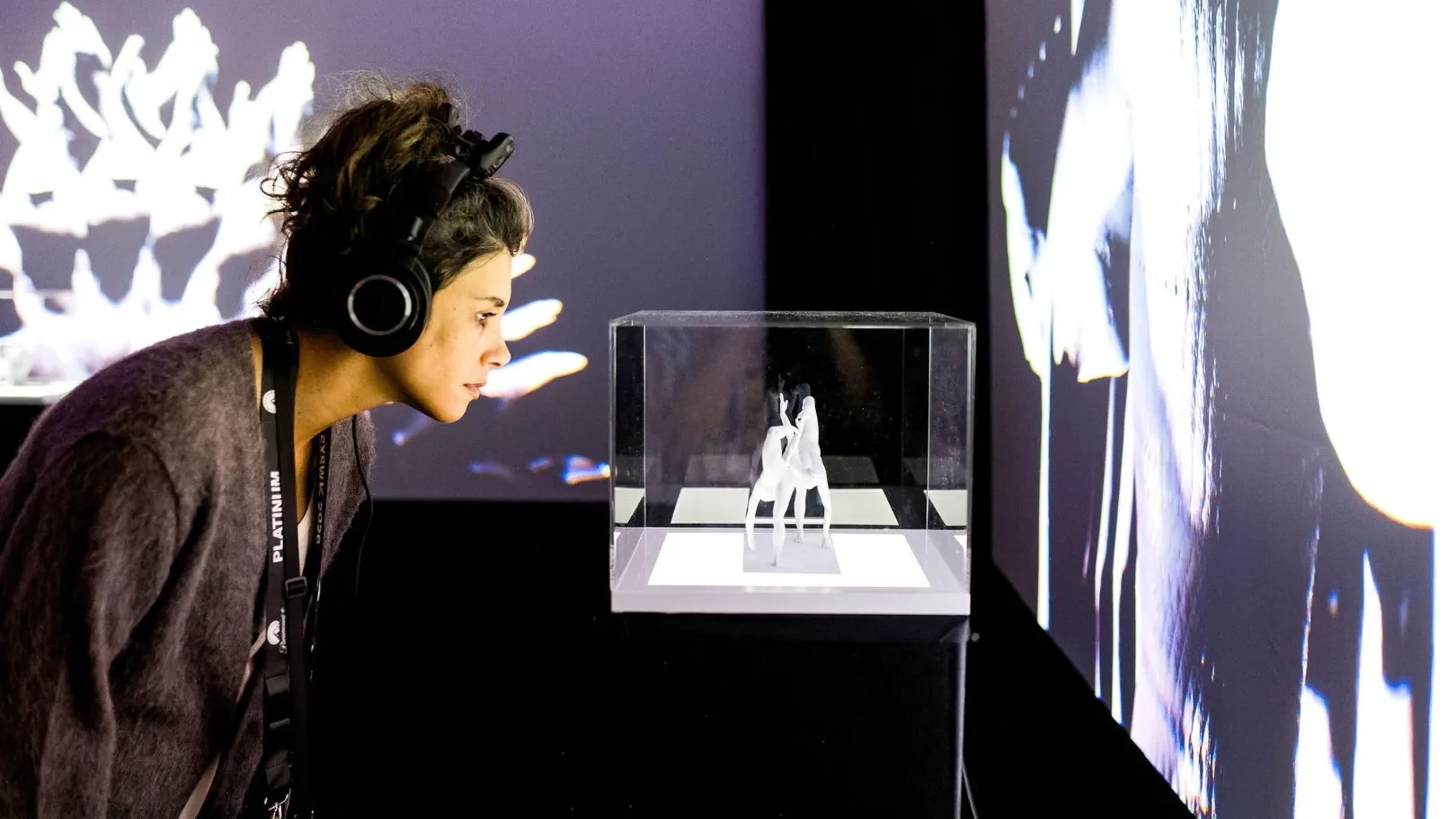 A women with headphone looking closely at a 3d printed sculpture of a female duet displayed in a vitrine srrounded by projections of a digital image of the photograpmetry of the duet