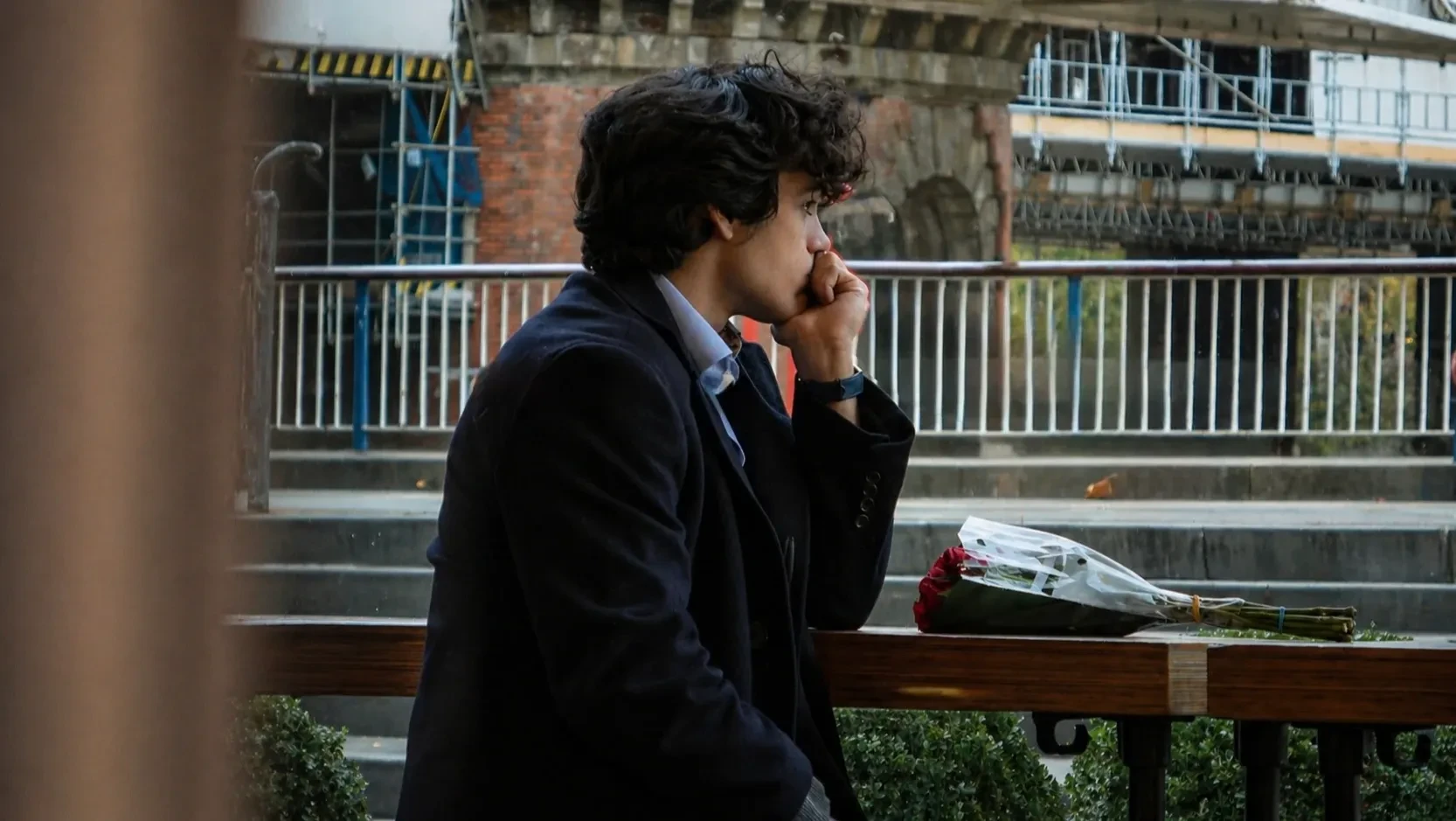A young man, sits alone in a cafe, deep in thought. A bouquet of disregarded roses in front of him.