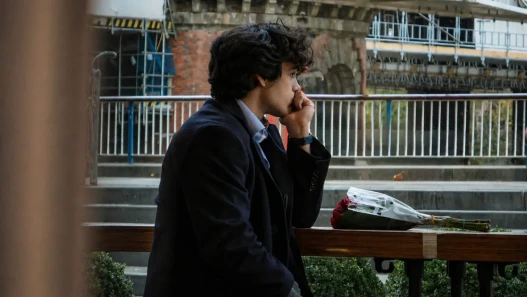 A young man, sits alone in a cafe, deep in thought. A bouquet of disregarded roses in front of him.