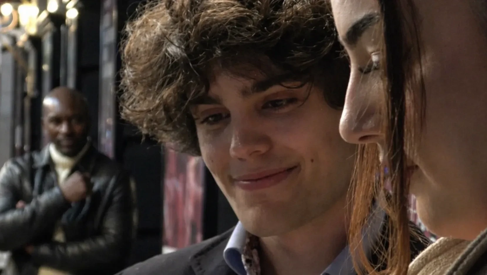 A young man and a young girl, stare at something together. Another man watches their interaction carefully from behind.