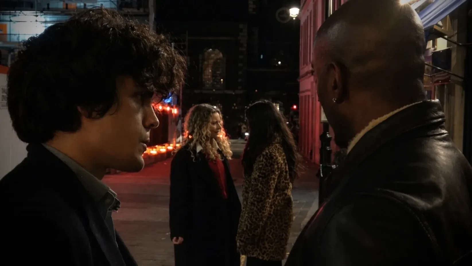 In a quiet street, a young man and an older man watch two girls having an intense conversation from afar.