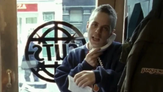 A man on a 1990's landline telephone in front of a shop window with logo 'CUTS' logo in the background