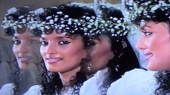 A young woman wearing a wreath of white flowers on her head looks directly at the camera through a distorted mirror showing three reflections.