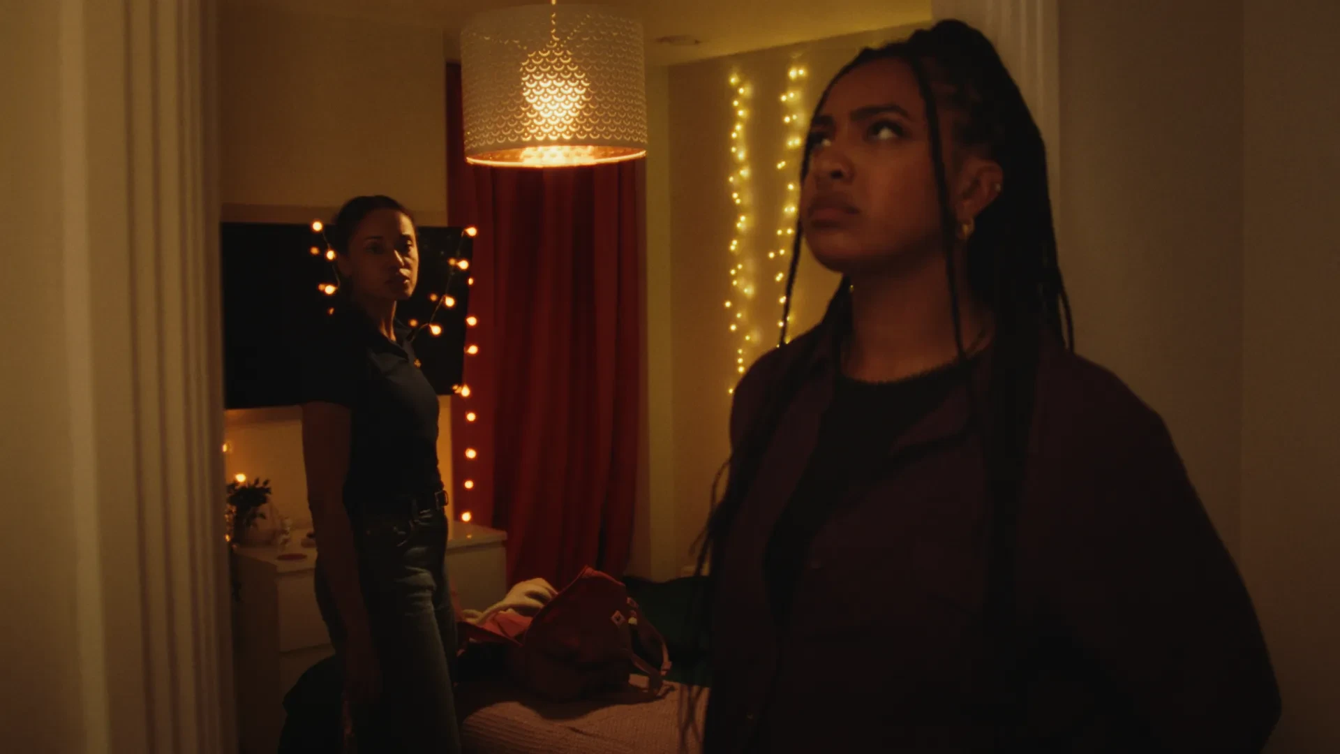 A woman leans against a doorframe, another woman looks on from within a bedroom.